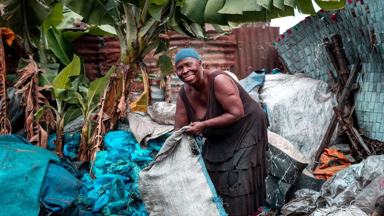 A woman stands outside next to bright green tree leaves. She holds a large sack containing plastic waste.