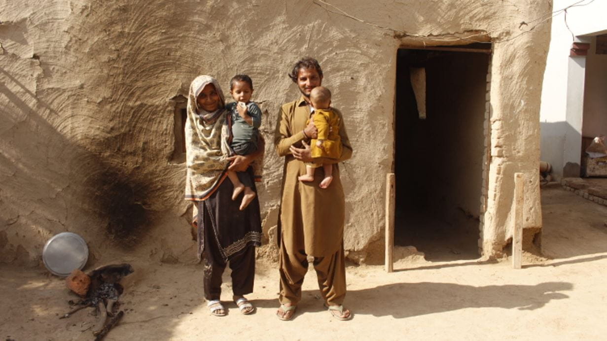 Mehru and her husband stand together outside their house. Each of the parents holds one of their young children.