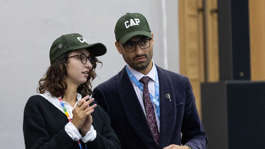 A man and a woman standing in conversation wearing green baseball caps with the word ‘CAP’ on them.