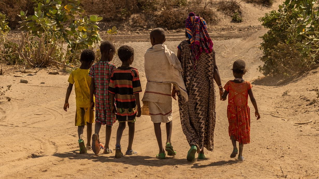 A woman and five of her children walking through a dried up riverbed with bushes in the background.