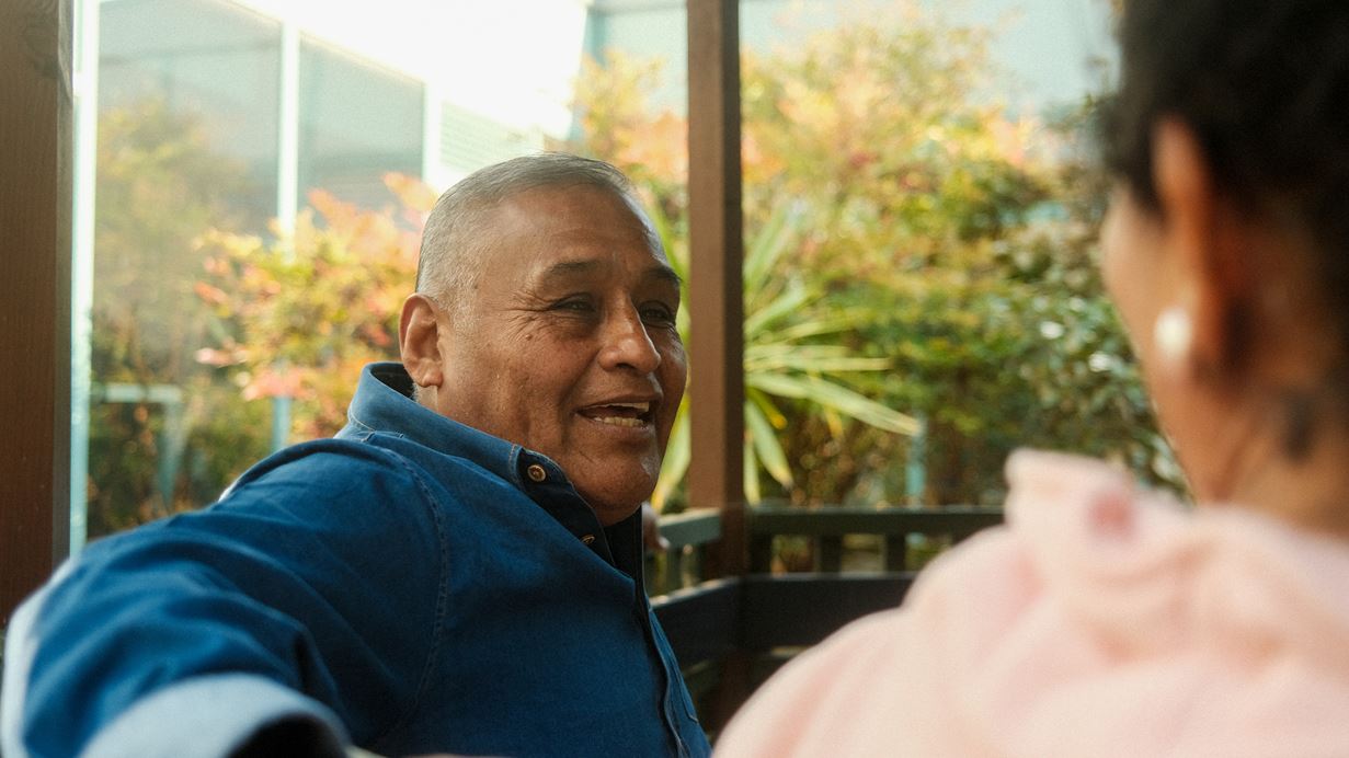 A South American man wearing a blue shirt talks to a woman in the backdrop of a garden.