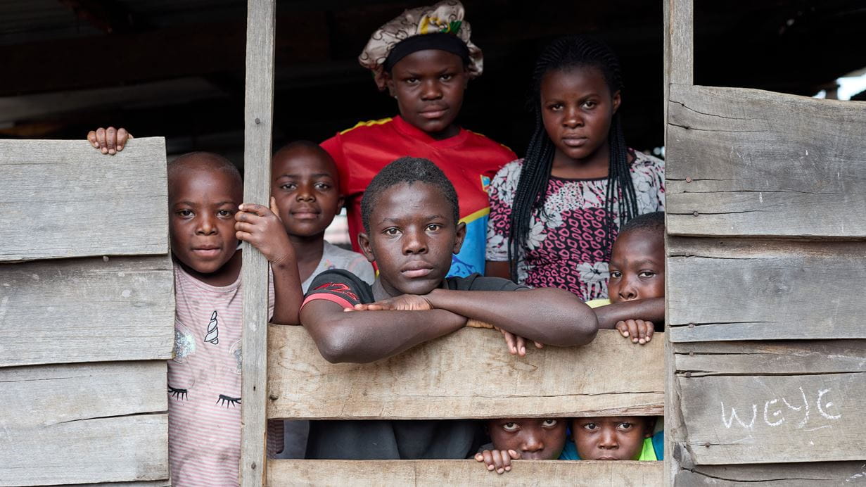 Seven children of various ages peer out between the planks that make up the wall of a classroom that has become their temporary home