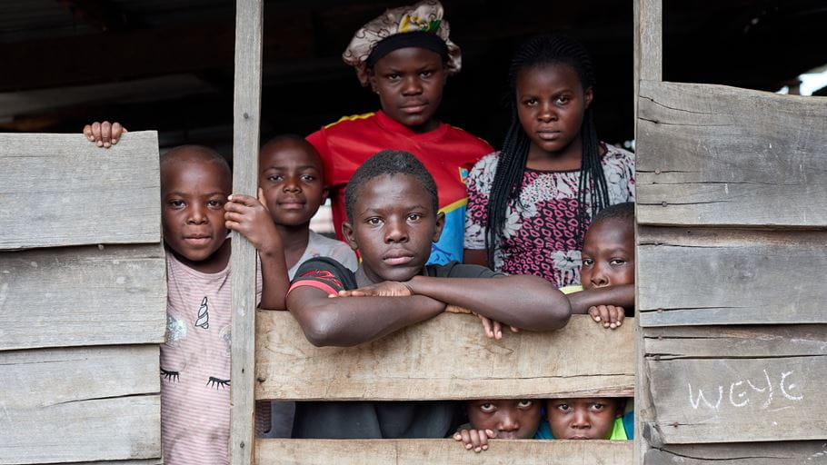 Seven children of various ages peer out between the planks that make up the wall of a classroom that has become their temporary home