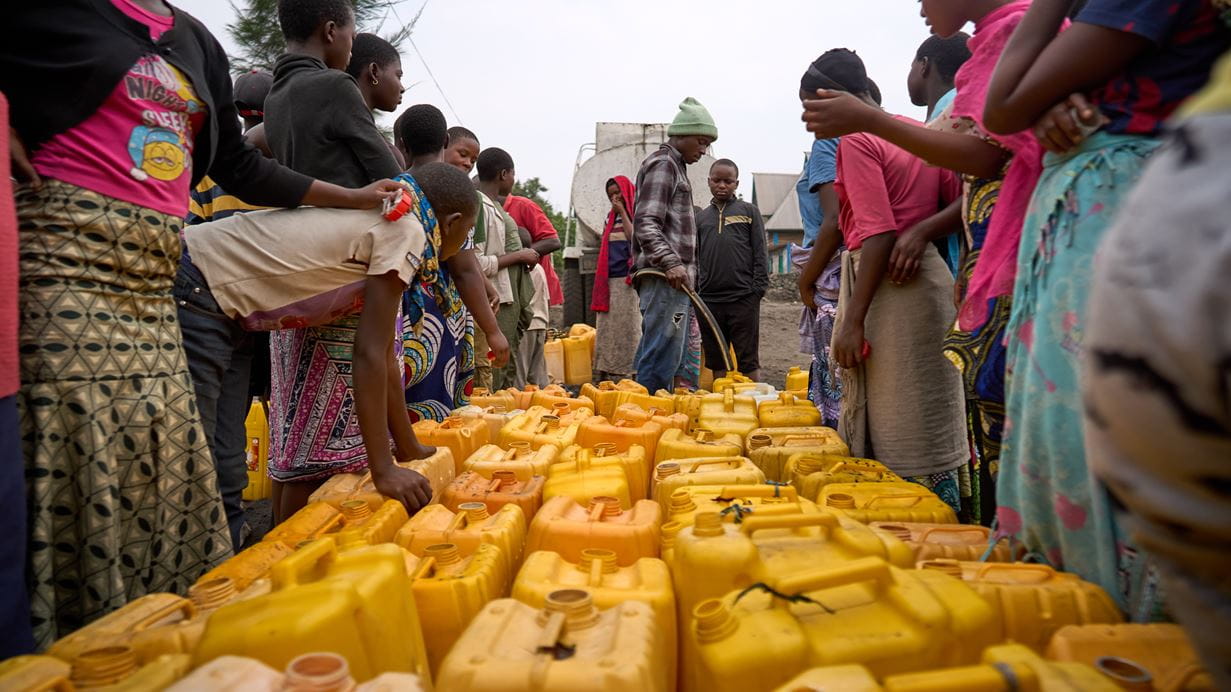 Many yellow jerry cans placed together ready to be filled with clean water while people wait alongside them