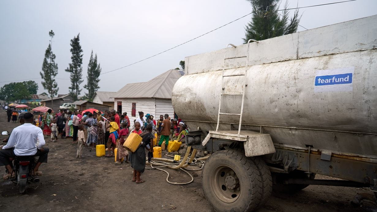 A large white truck with a blue Tearfund logo carries clean water for people waiting 