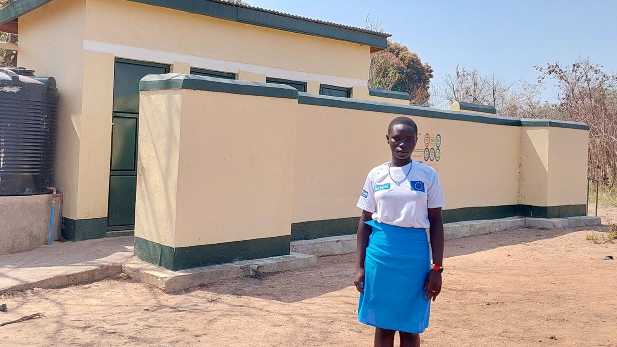 A girl wearing a white tshirt with the Tearfund logo on, stands in front of a small building.