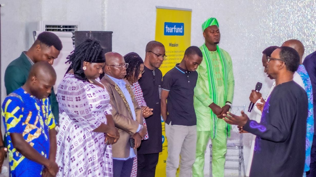 A group of people stand together with their eyes closed as someone with a mic in their hand prays. Behind them is a yellow Tearfund banner with the logo at the top.