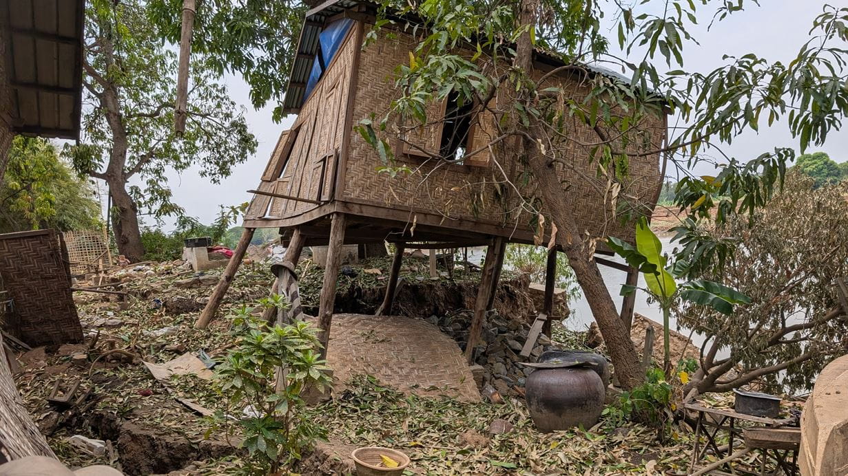 A wooden house on stilts lists to one side after being damaged by the 28 March earthquake in Myanmar. It is surrounded by green vegetation.