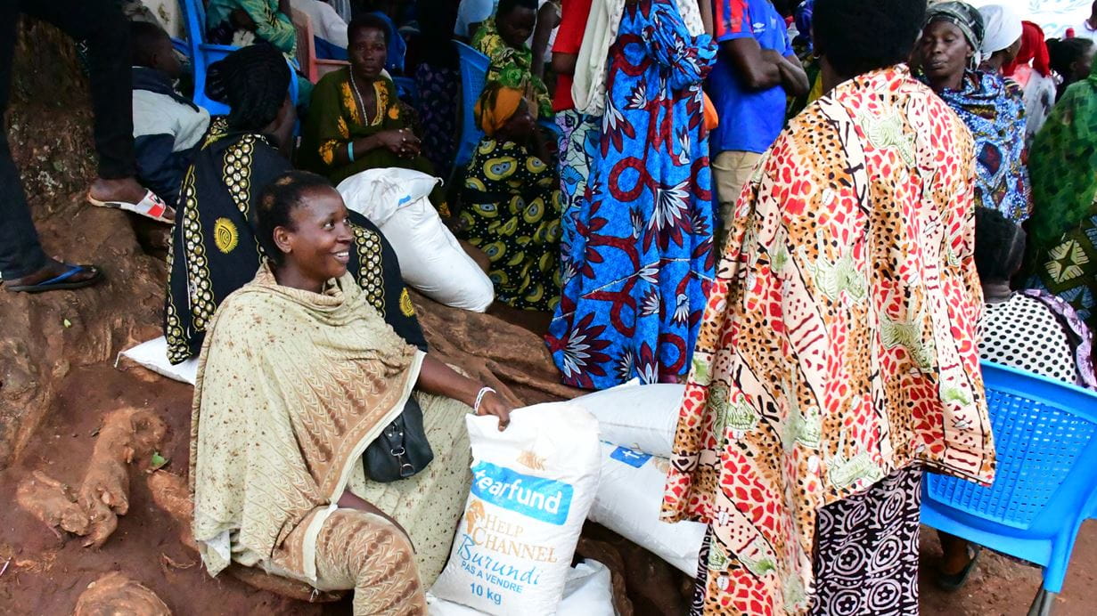 A woman sits amongst a group of woman standing nearby. She smiles as she shows another woman the bag of food she has been given.