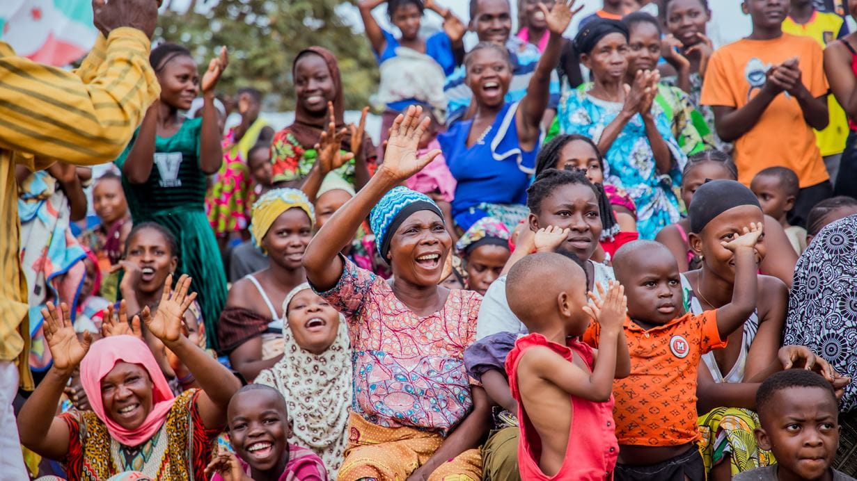 A group of people, mostly women and children, sit in a group outdoors under a bleu sky. They are dressed in a variety of bright colours. Many of them are smiling and raising their hands in the air in a happy gesture.