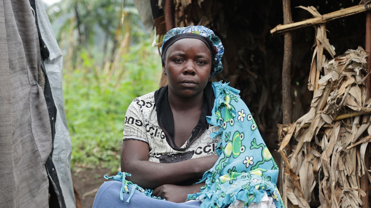 A woman sits facing the camera in front of a makeshift shelter
