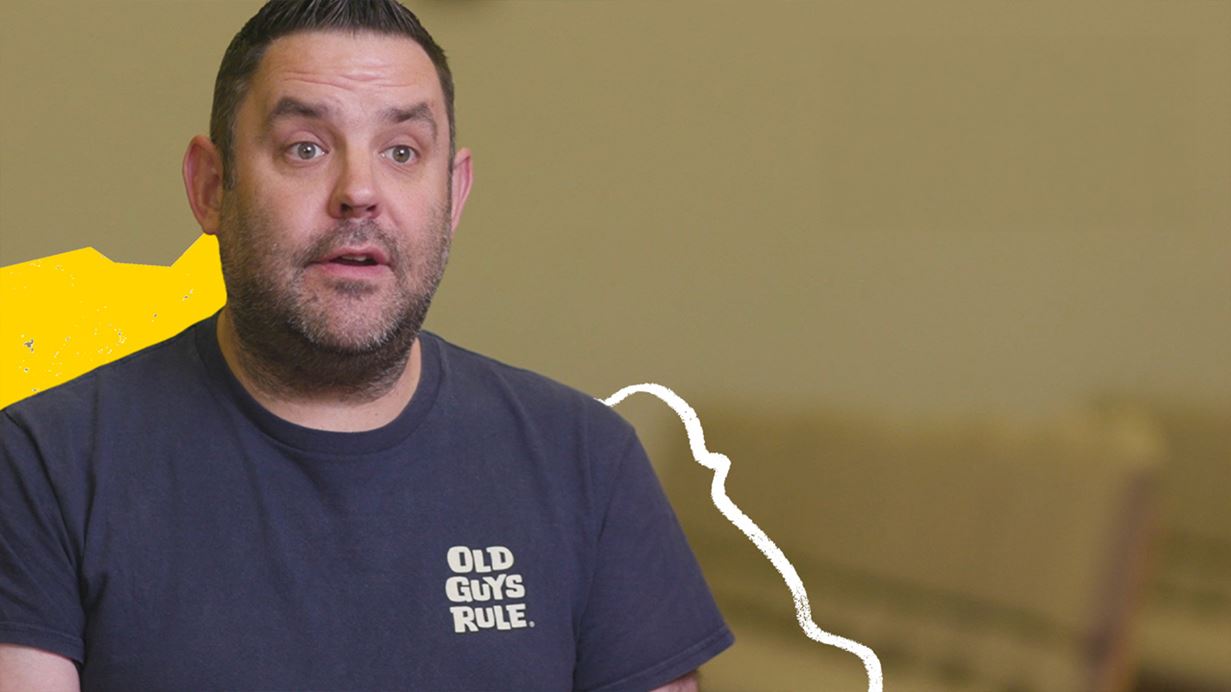 A man wearing a dark blue tshirt speaks to the camera