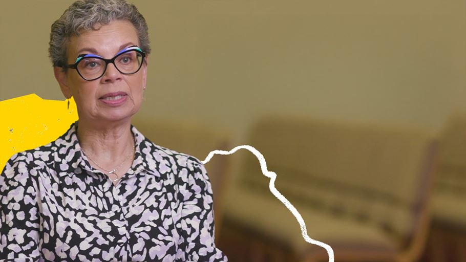 A woman wearing glasses with short grey hair speaks to the camera. 