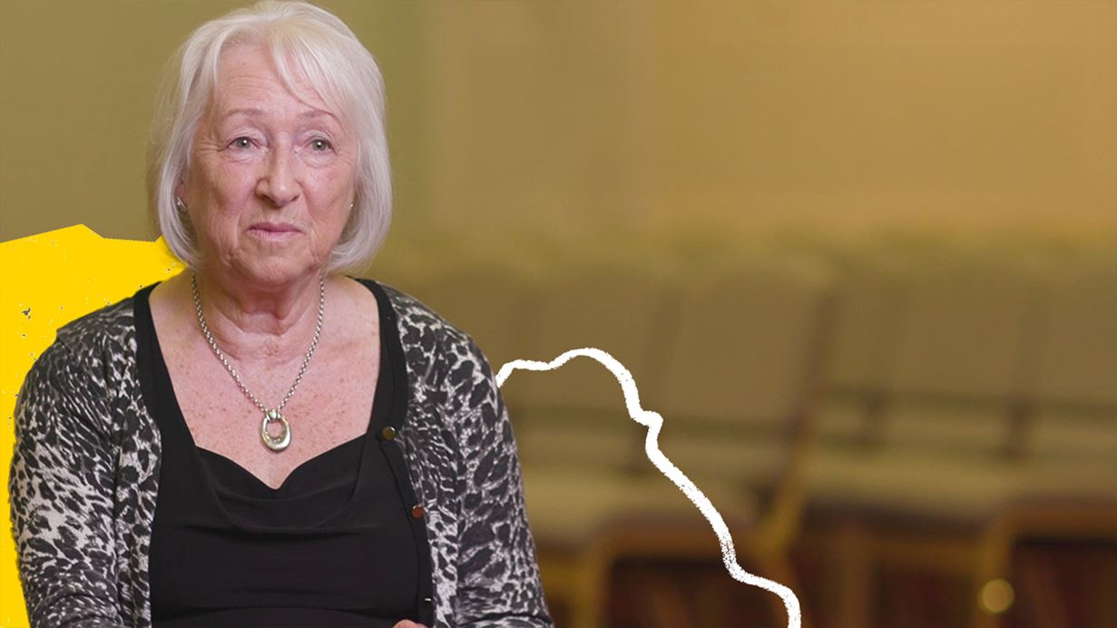 A woman with short grey hair speaks to the camera. 