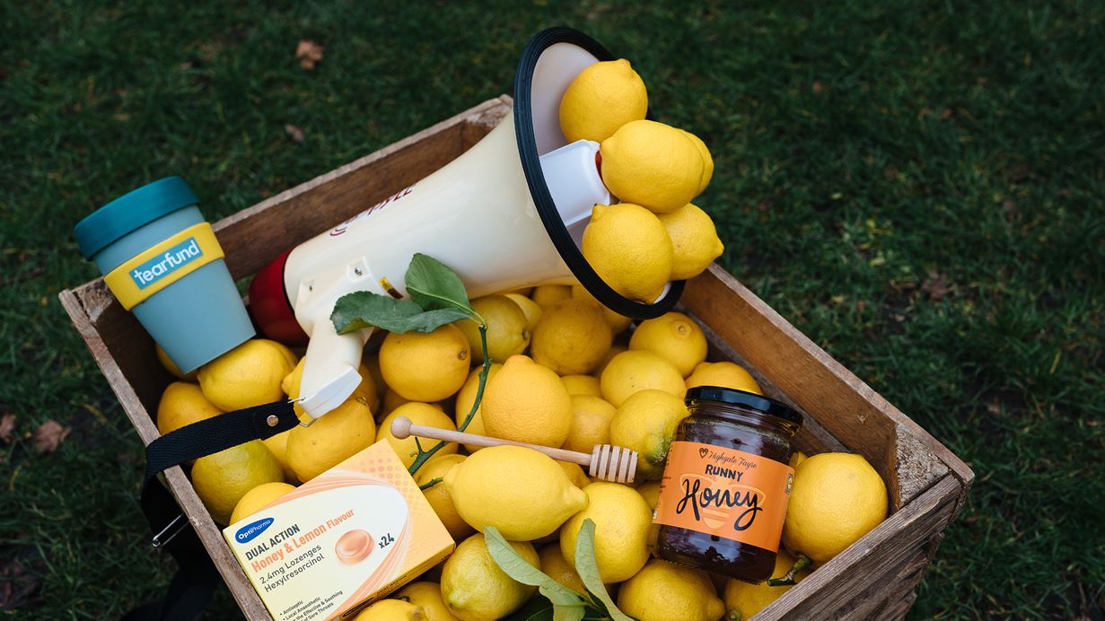 On top of a crate filled with lemons are a Tearfund keep cup, a speaker, a jar of honey, a wooden dipper and lozenges.