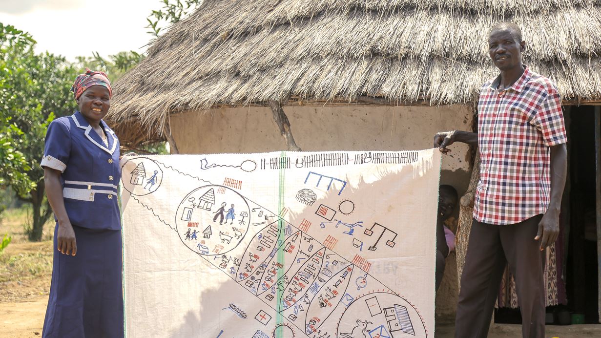 John and Helen stand outside their house with a thatched roof, holding between them the life map that they created