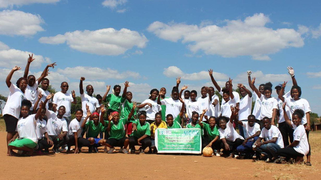 A group of people outside under a blue sky wearing MCCN T-shirts and displaying a banner speaking about platsic waste