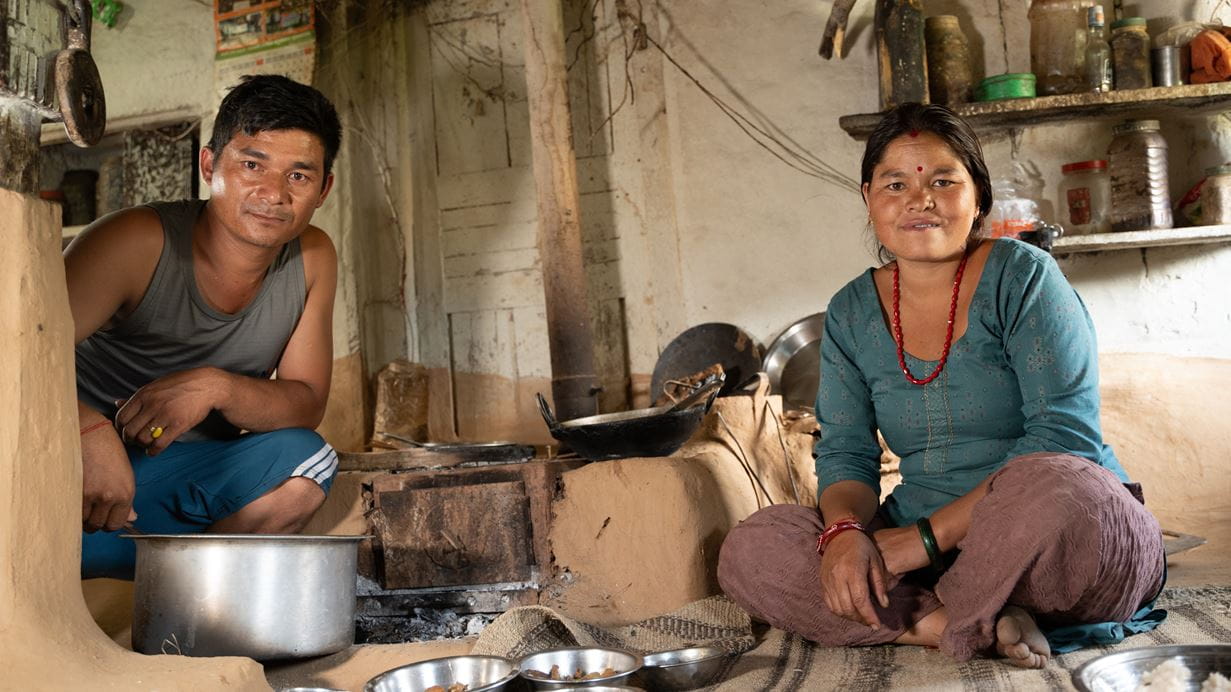 A man and a woman in traditional Nepali dress sit inside their home with bowls of food