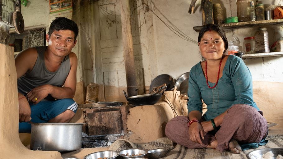 A man and a woman in traditional Nepali dress sit inside their home with bowls of food
