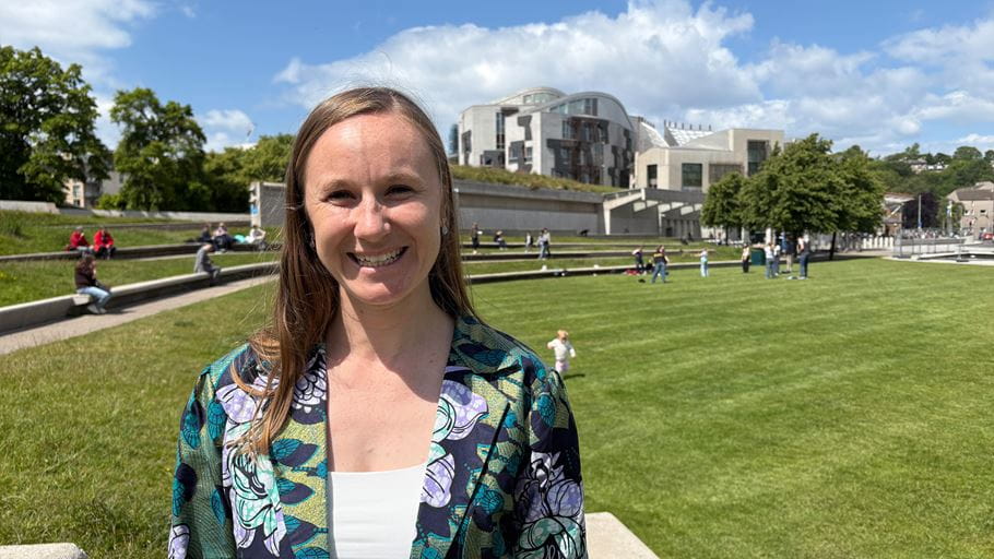 A woman, Poppy, smiles. Stood in a large green space in front of the the Scottish Parliament building.