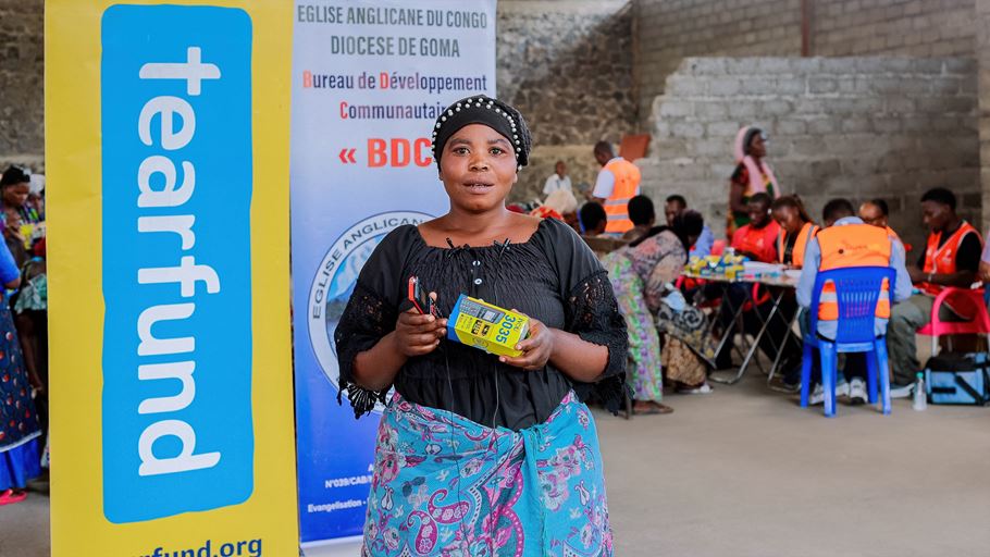 A woman holds a phone and its box in her hands. She stands in front of a Tearfund banner and several people sat are tables in the background.