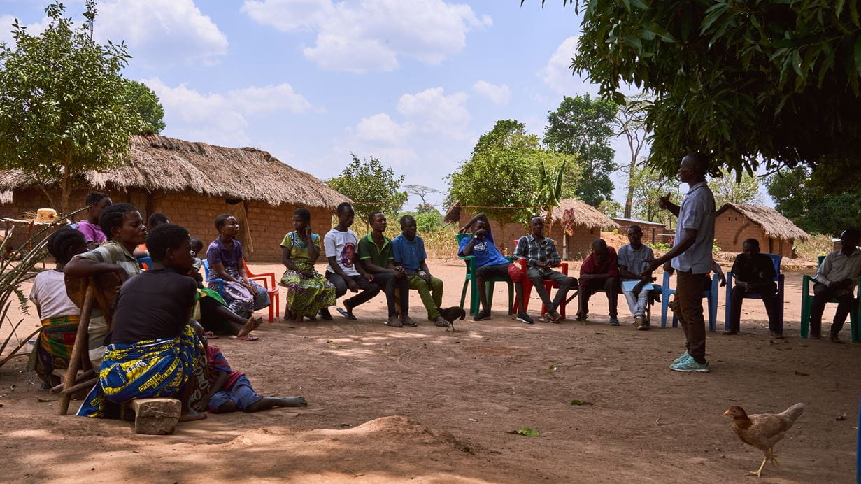 A group of men and women sit on chairs in a circle outside in the shade of a large tree while one man speaks to them about peacebuilding.