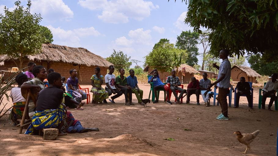 A group of men and women sit on chairs in a circle outside in the shade of a large tree while one man speaks to them about peacebuilding.