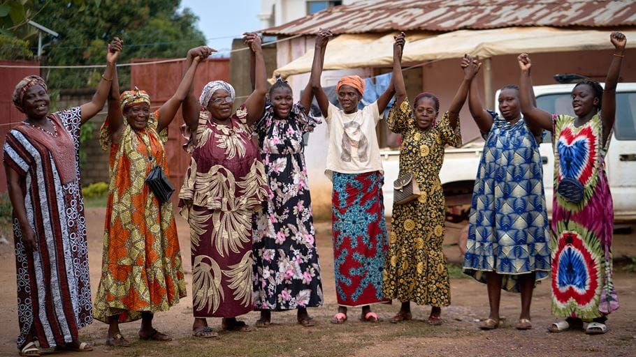 A group of African women holding hands with arms raised, wearing colorful traditional dresses.
