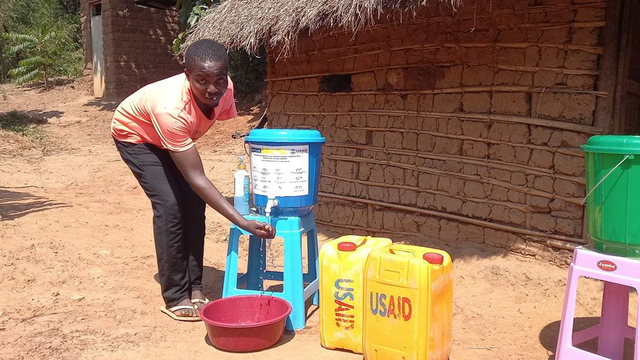 Man washes hands at a simple water station in rural setting with mud hut background.