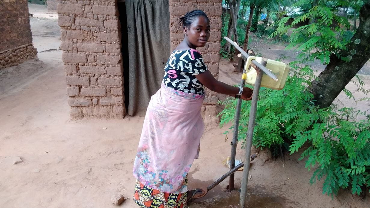 Woman washing hands at rural handwashing station near a mud brick building.