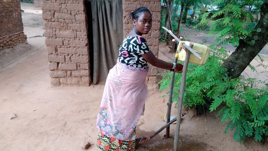 Woman washing hands at rural handwashing station near a mud brick building.