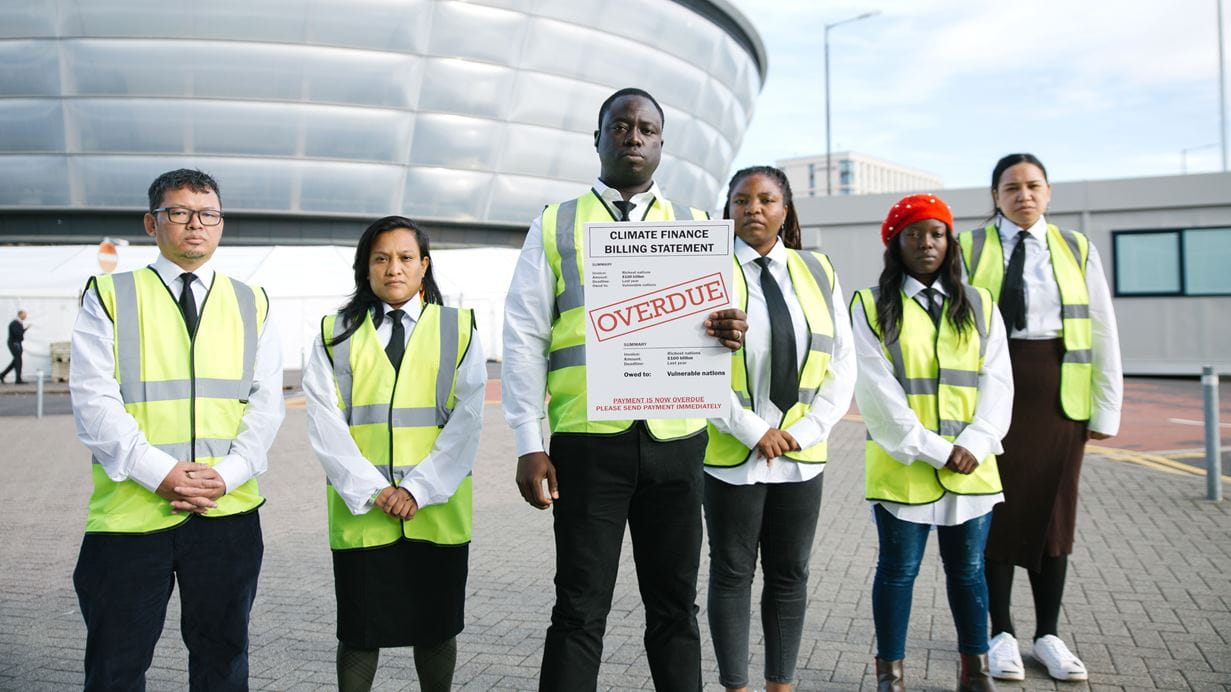 Group of activists holding an "Overdue" climate finance billing statement, wearing hi-vis vests.