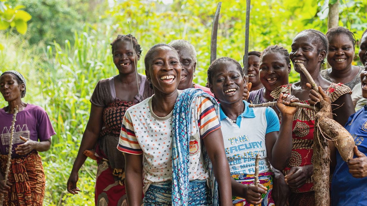 A group of smiling African women stand together in a lush, green, rural setting.