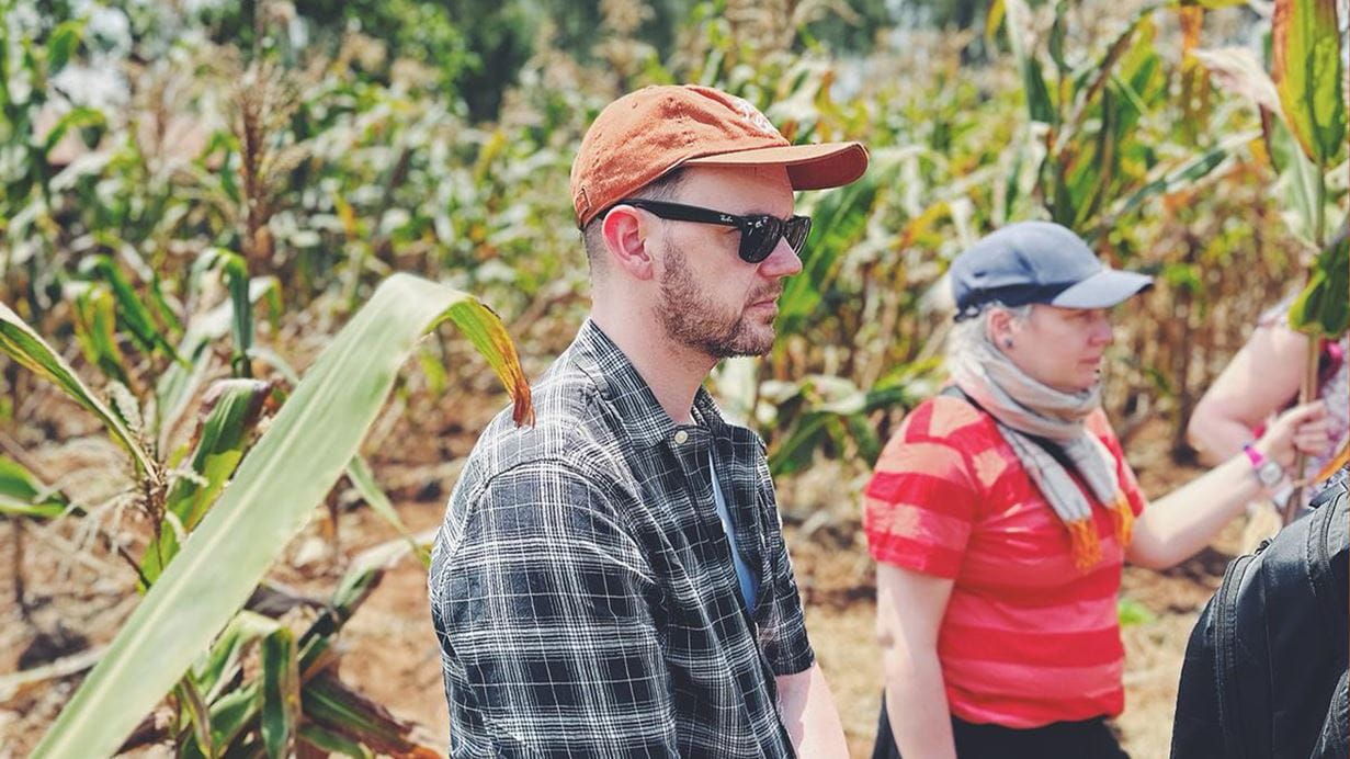 People in a corn field, one in an orange hat, the other in a blue cap, both wearing sunglasses.