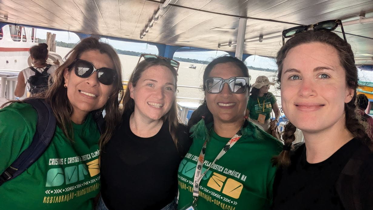 Four smiling women pose on a boat deck, some wearing climate justice t-shirts. River view behind.