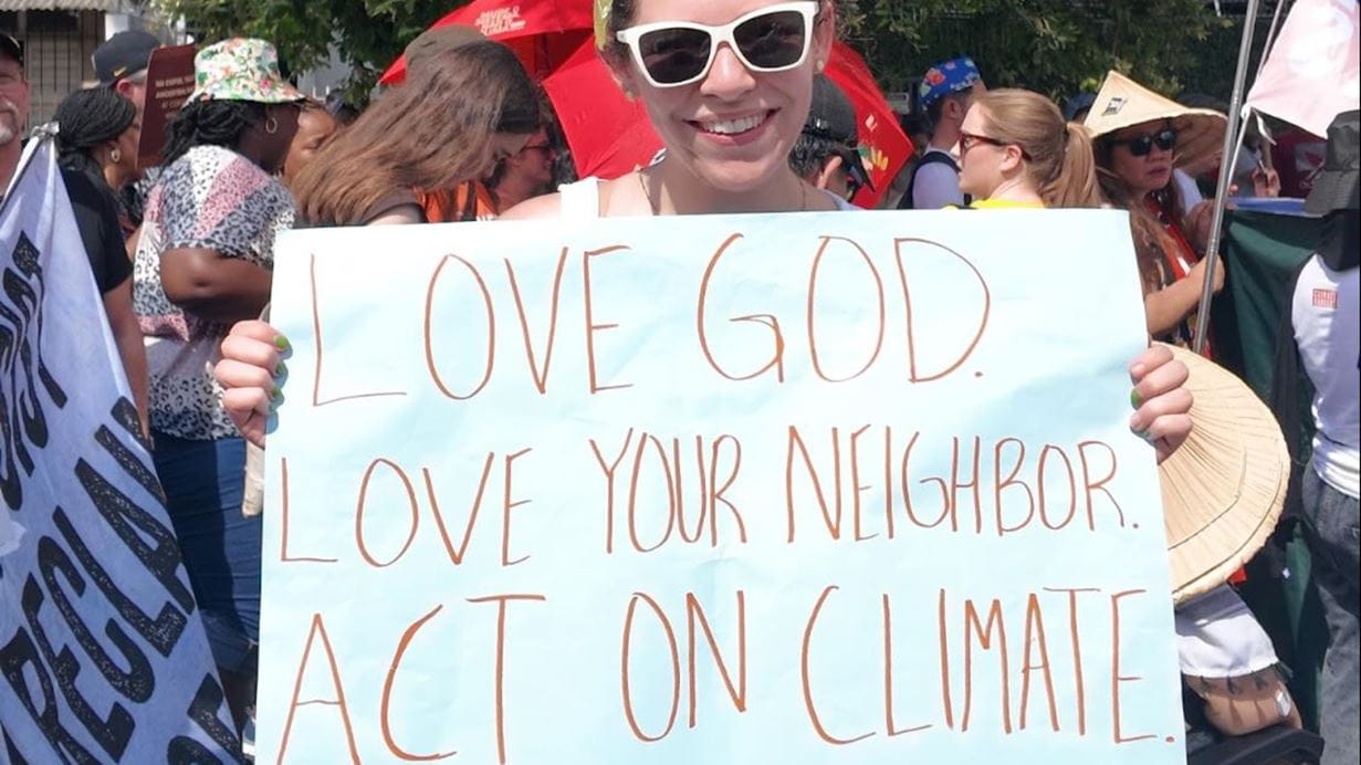 Smiling woman at a climate rally holds sign reading "Love God. Love Your Neighbor. Act on Climate."