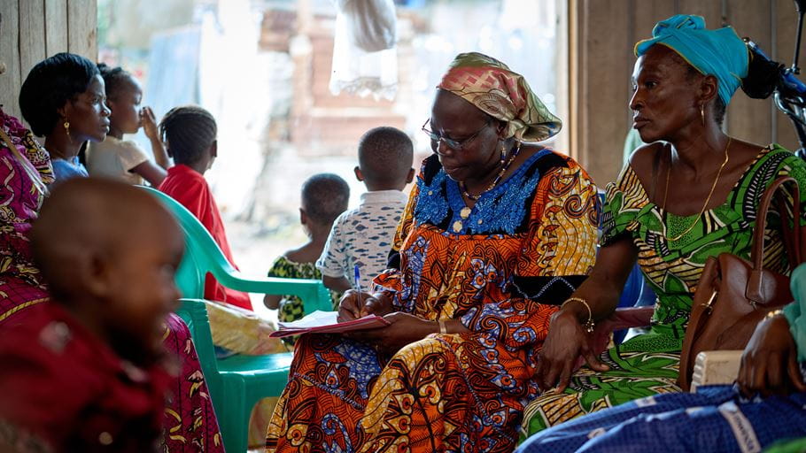 Photograph shows women in colorful African dresses, one writing in a notebook, with children in background.