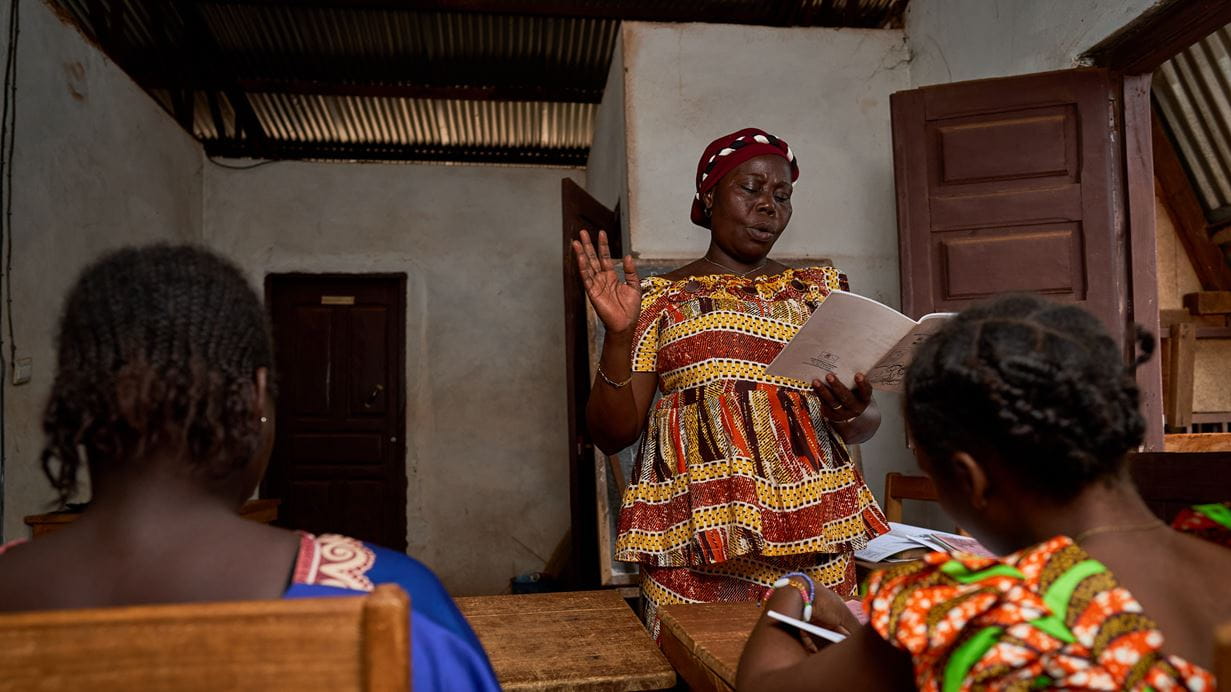 African woman in patterned dress leads classroom discussion with students at desks.