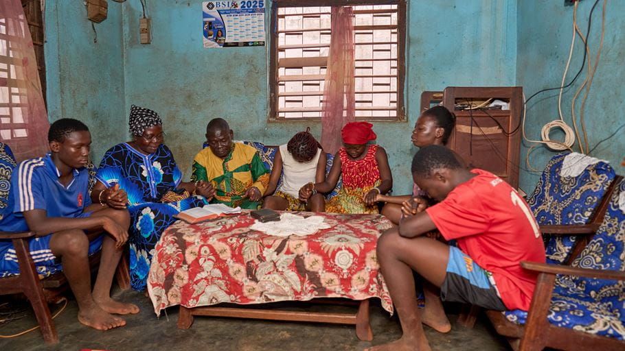 Family gathered indoors praying together around a table in a modest home, faith and tradition.