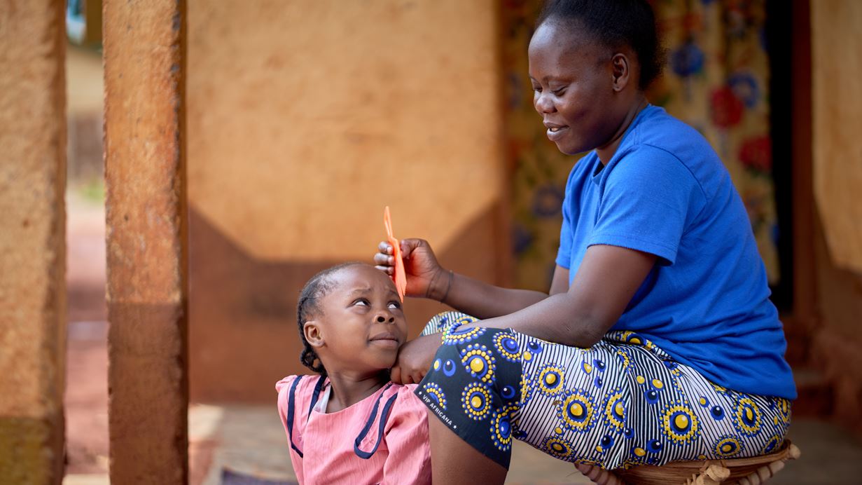 A woman combs a young girl's hair outside a home. Both are smiling and the girl is looking up.
