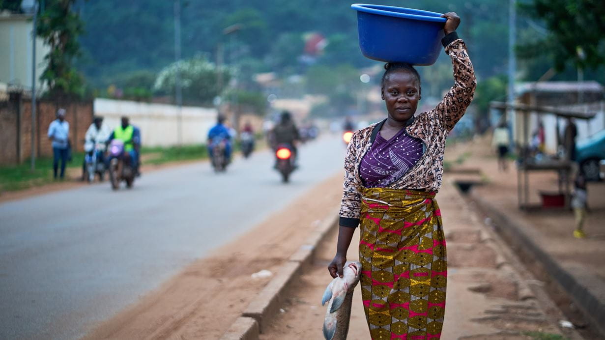 Woman carrying a blue basin on her head and holding fish on a road in an African village.