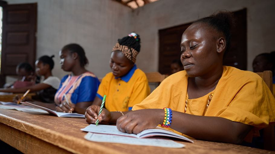 African women in classroom, writing in notebooks on wooden desks, wearing colorful clothes.