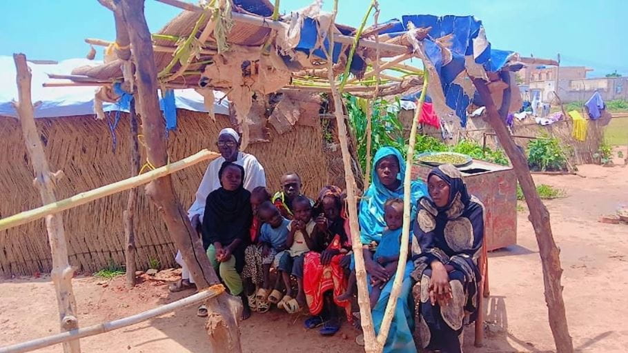 Family gathered under makeshift shelter in arid landscape, likely displaced or in a developing area.