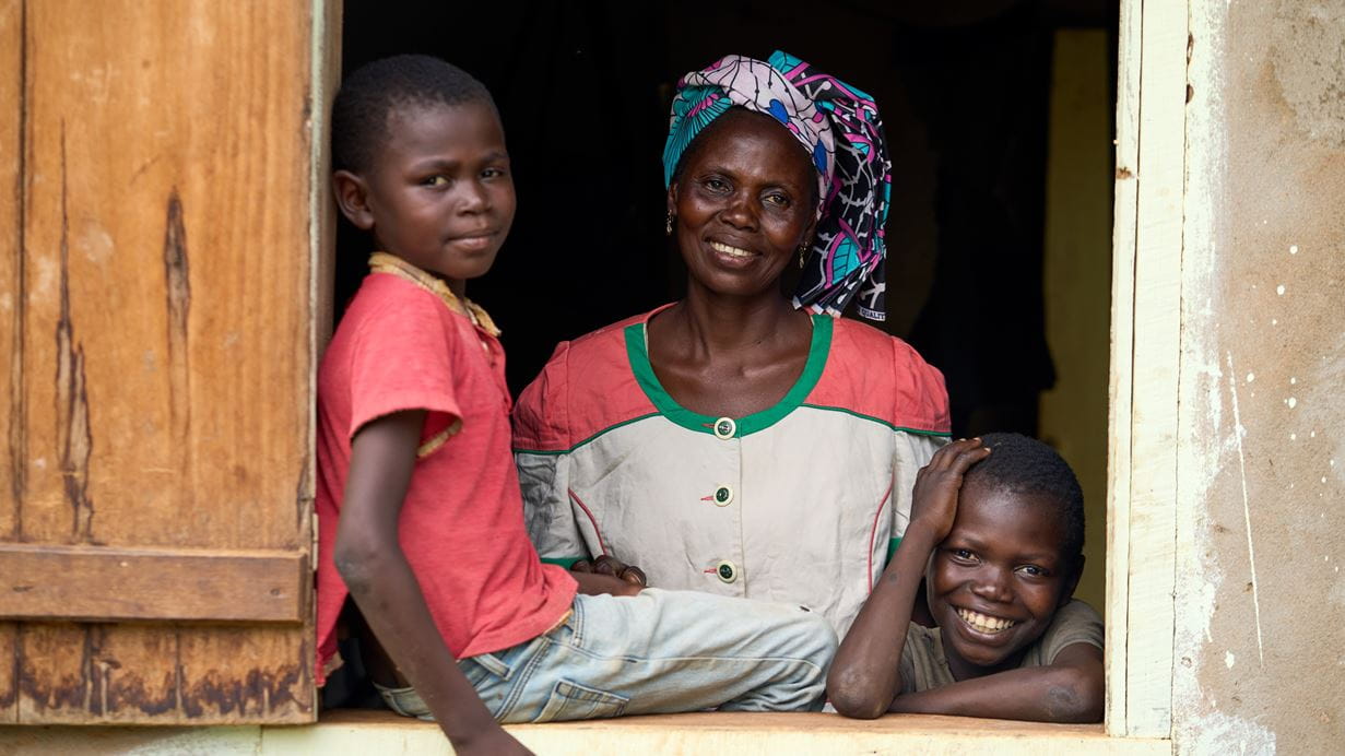 A woman in a colourful headscarf and her two young sons look through a window and smile brightly at the camera