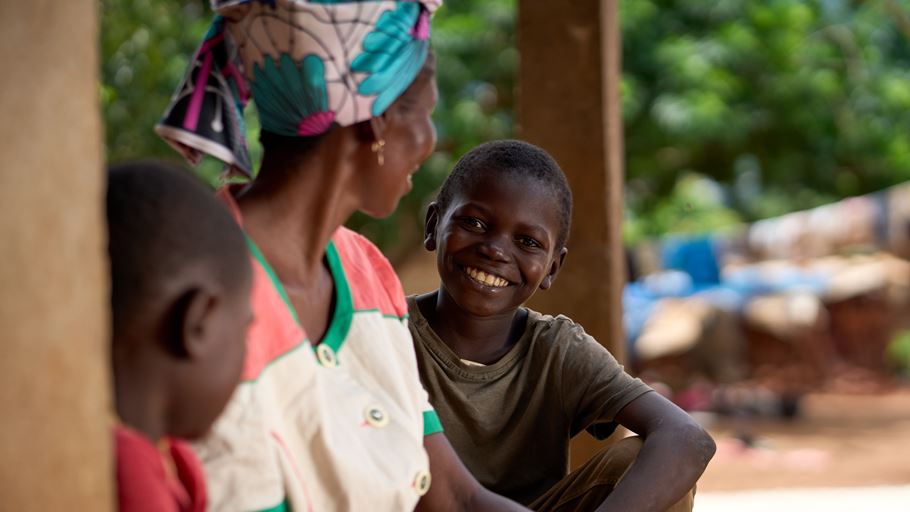 A smiling African boy sits with family, the woman wearing a colorful patterned headscarf.