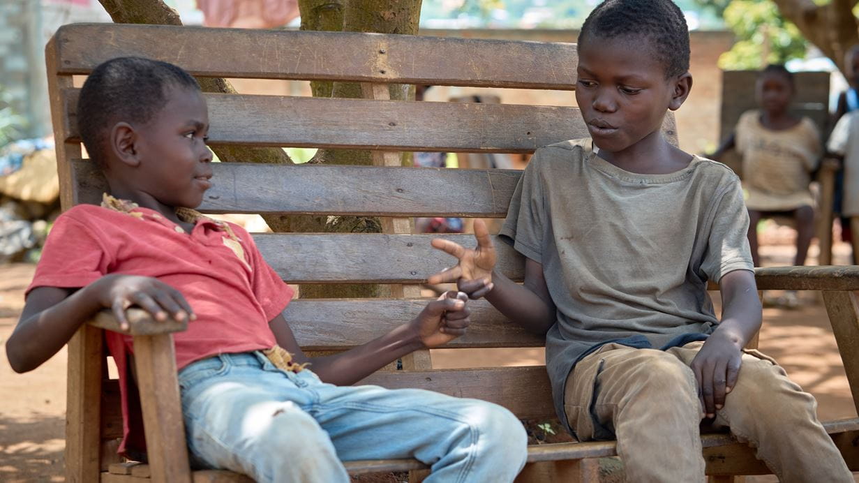 Two African children play a game on a wooden bench in a village, with other children nearby.