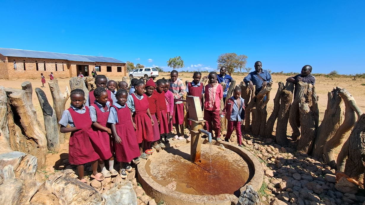 Group of children in red uniforms stand around a water pump in a rural African village.