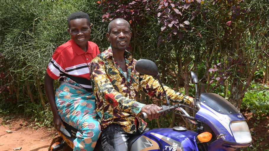 A couple smiles from a motorcycle. The man sits in front, woman behind, bushes in the background.