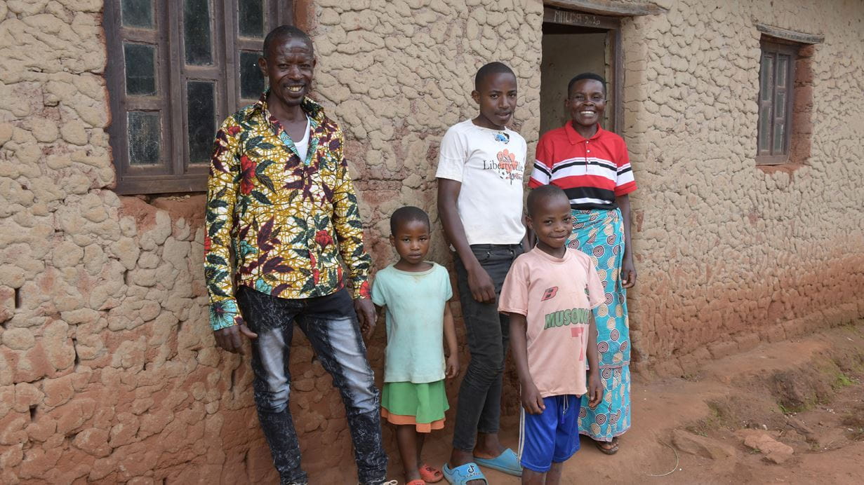 A family of five poses in front of a textured mud house with windows and a doorway.