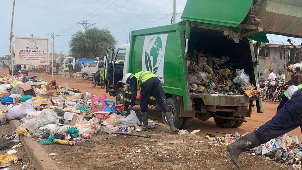 A street in Juba, South Sudan, where two EcoClean employees are gathering loose waste strewn across the ground and loading it into their green truck.